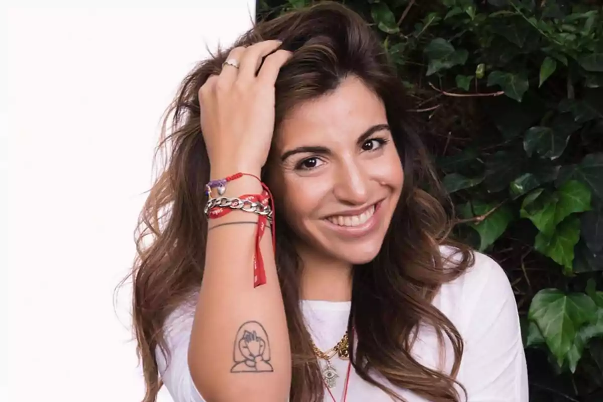 Mujer sonriente con cabello largo y castaño, usando pulseras y mostrando un tatuaje en el antebrazo, posando frente a un fondo de hojas verdes