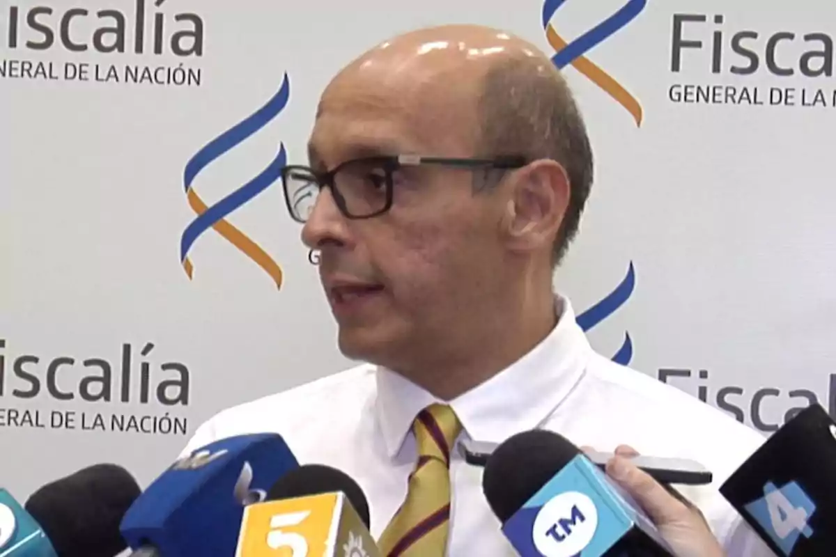 Man with glasses and a tie speaking in front of several microphones at a press conference with the Attorney General’s Office logo in the background Man with glasses and a tie speaking in front of several microphones at a press conference with the Attorney General’s Office logo in the background
