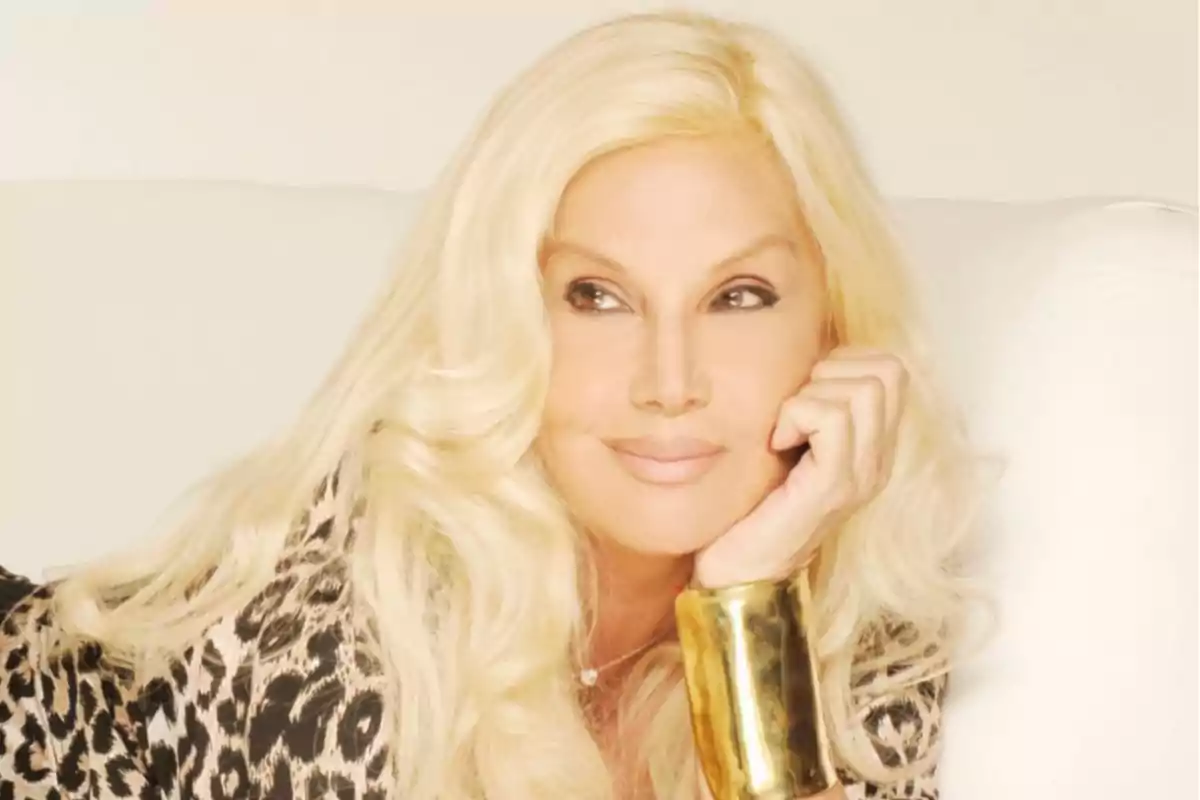 Mujer rubia con cabello largo y ondulado, apoyando su rostro en una mano, usando un brazalete dorado y una prenda con estampado animal print, mirando hacia un costado con expresión relajada