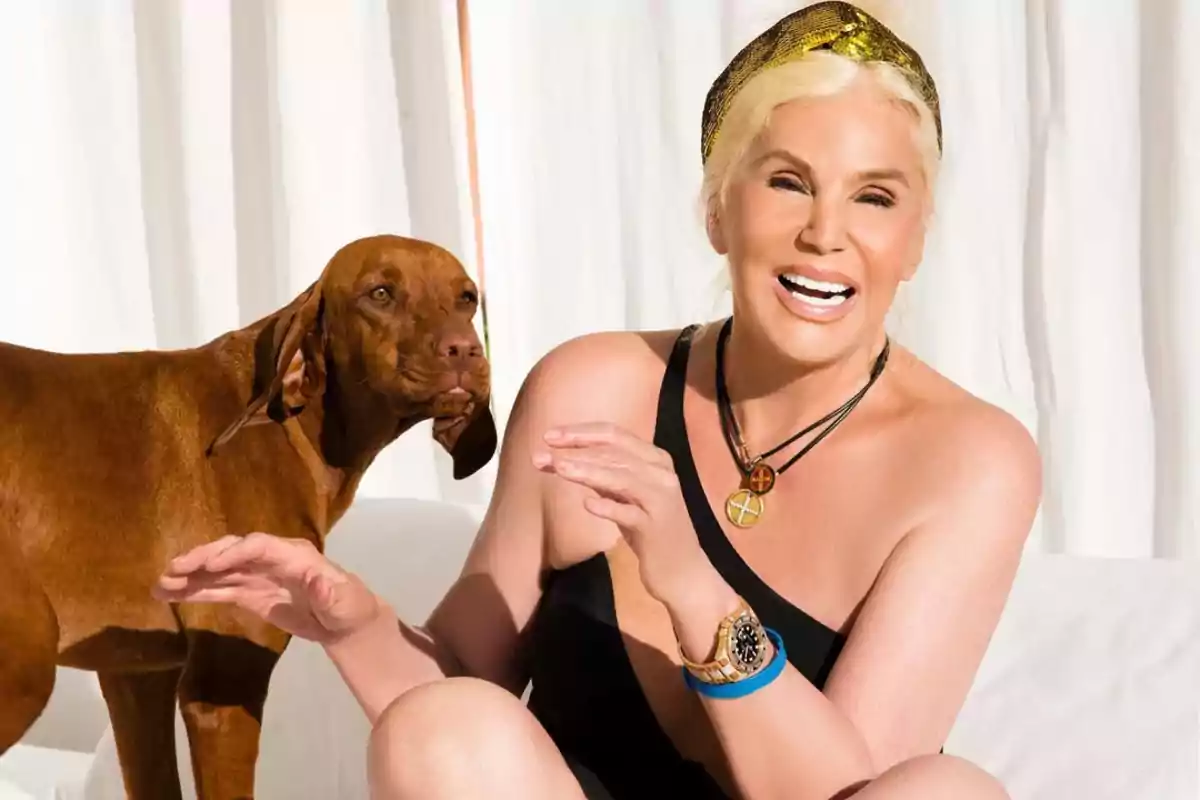Smiling blonde woman in a black dress with a gold headband sitting next to a brown dog in front of a white curtain