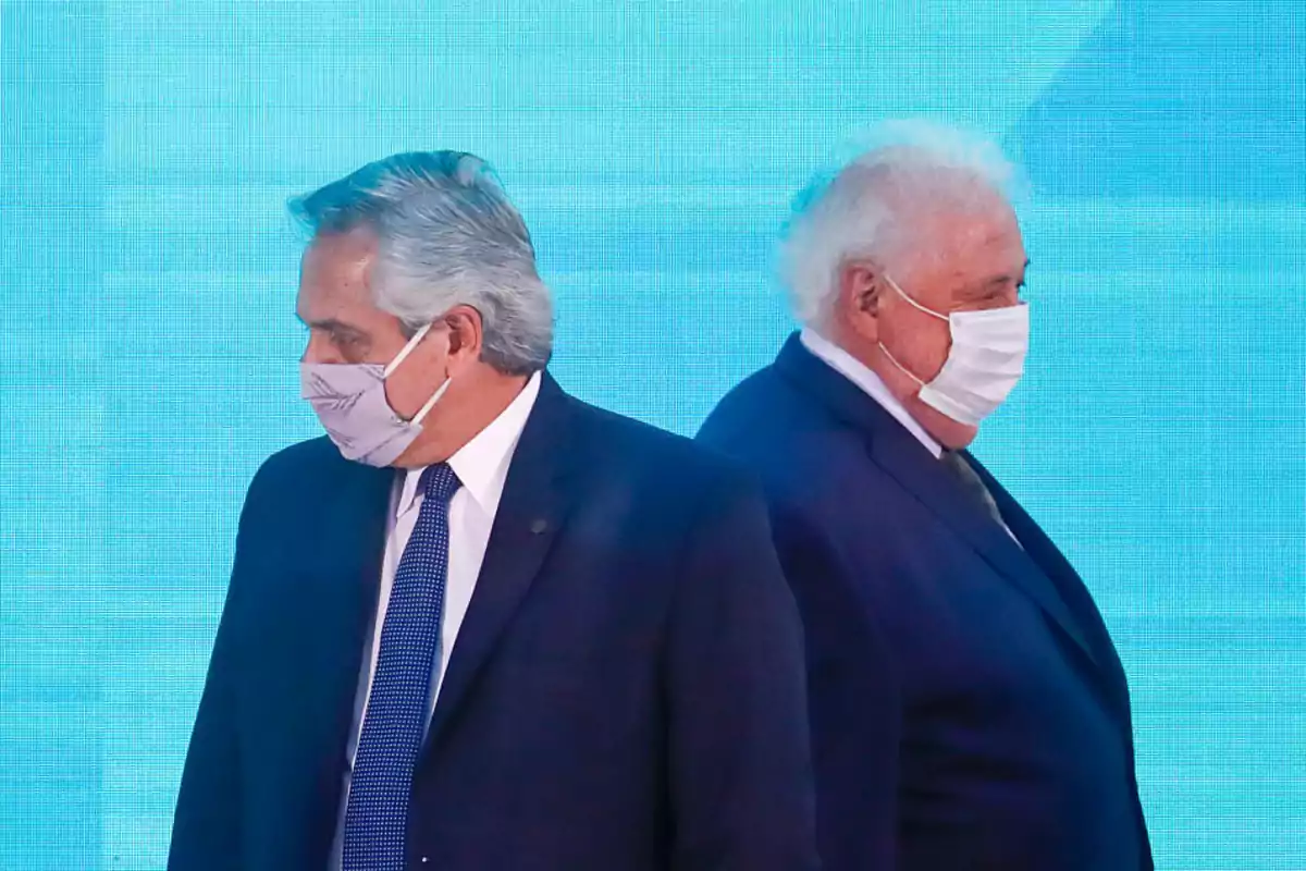 Dos hombres de traje y mascarilla miran en direcciones opuestas frente a un fondo azul.