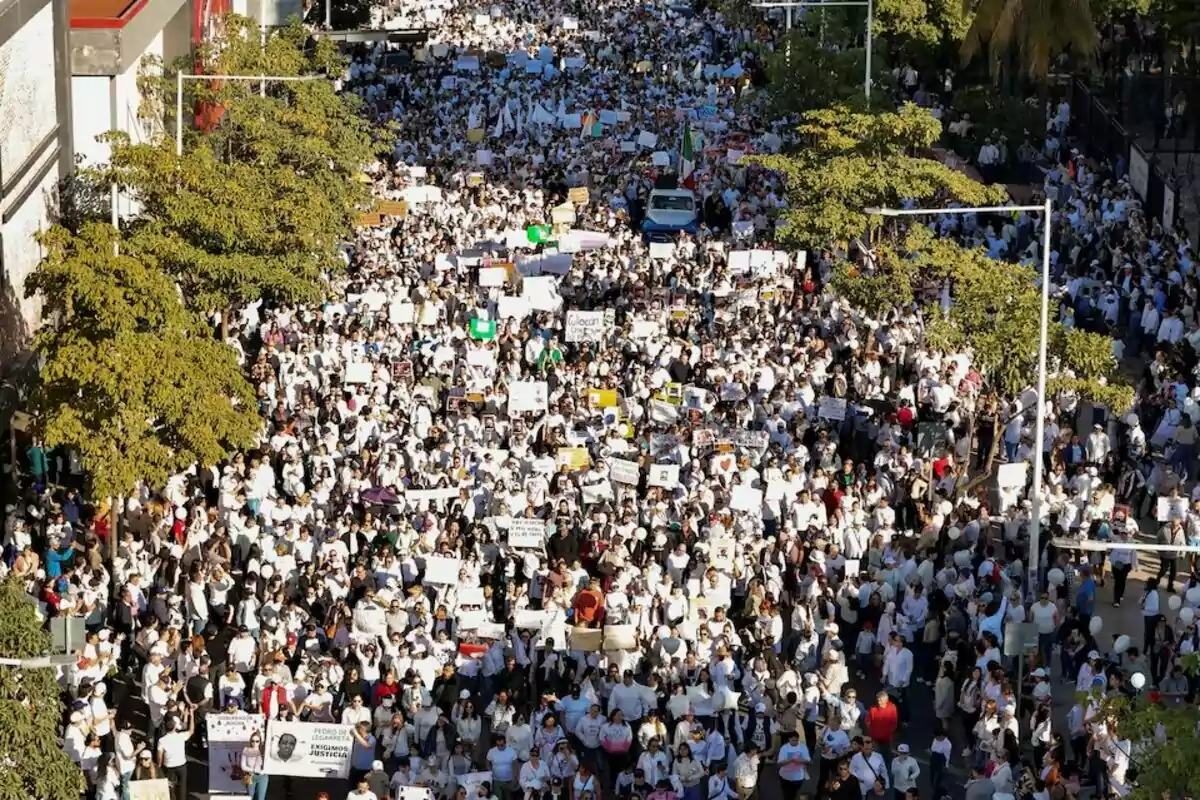 Una multitud de personas vestidas de blanco participa en una marcha pacífica en una calle arbolada, sosteniendo pancartas y carteles.