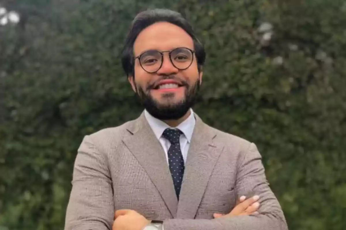 Young man with a beard and glasses smiling, wearing a beige blazer and a dark tie, arms crossed in front of a background of greenery. Young man with a beard and glasses smiling, wearing a beige blazer and a dark tie, arms crossed in front of a background of greenery.