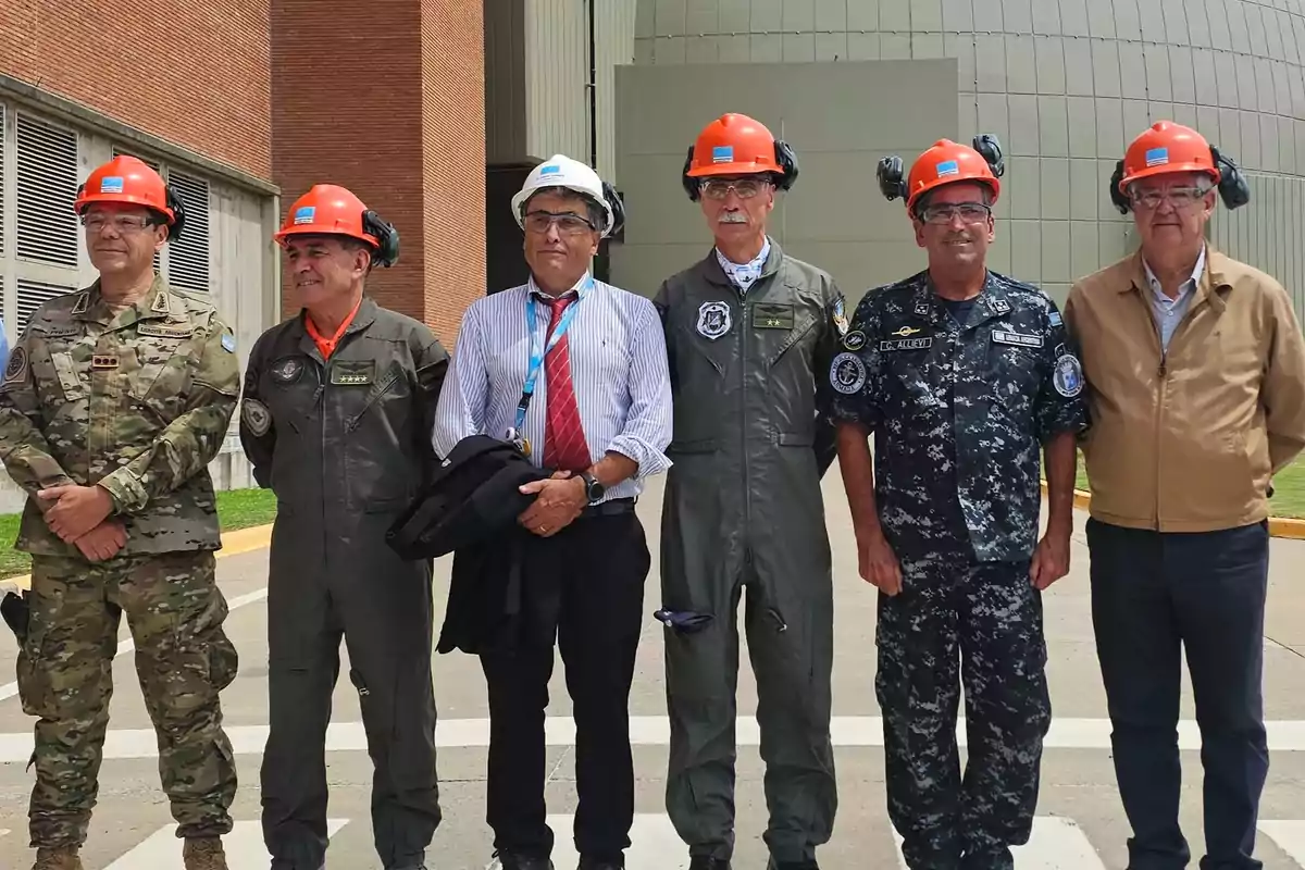 Un grupo de seis hombres con cascos de seguridad y uniformes militares posan frente a un edificio industrial.