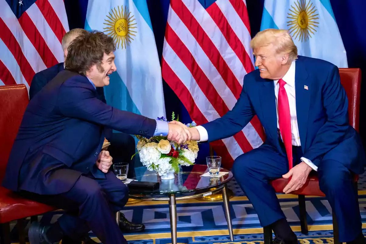 Two people shaking hands while sitting at a table with United States and Argentina flags in the background.