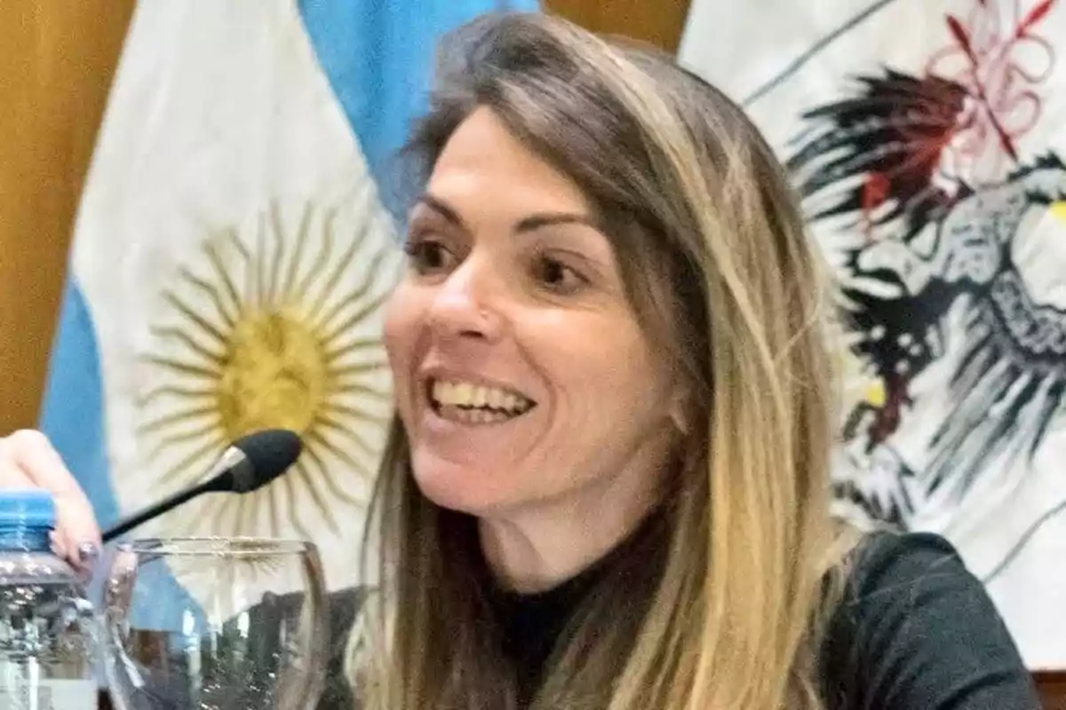 A smiling woman speaking into a microphone with flags in the background.