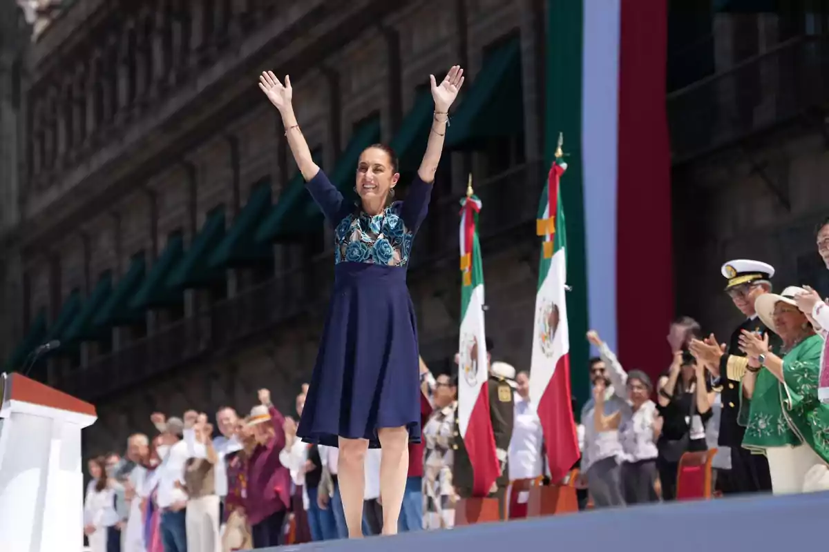 Una persona con los brazos levantados en un evento público frente a una multitud y banderas.