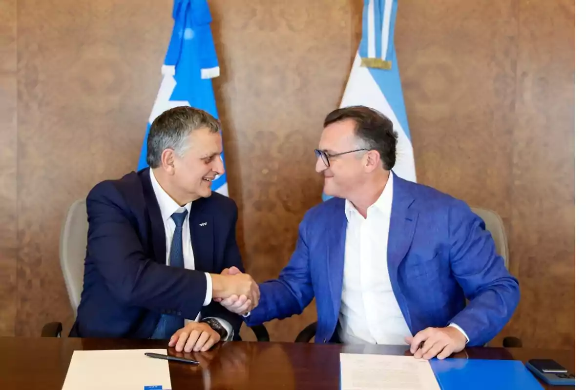 Two men in suits shake hands at a table with documents and flags in the background.