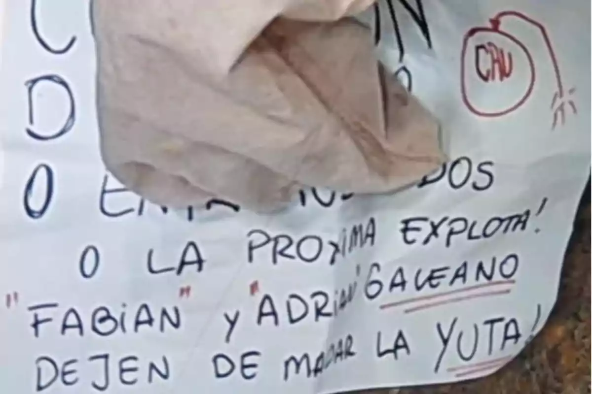 Un papel con un mensaje escrito a mano, parcialmente cubierto por un objeto marrón, con un dibujo de una bomba y texto en letras mayúsculas.