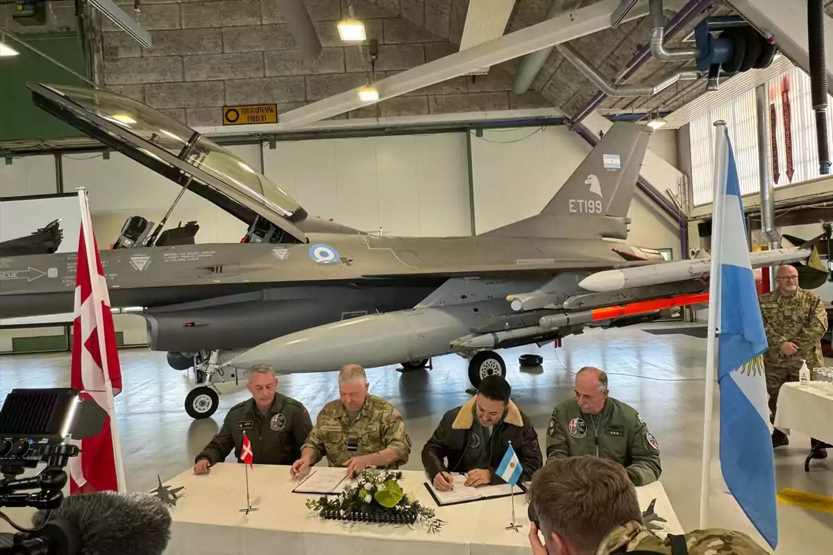 Militares de Argentina y Dinamarca firman documentos en una mesa frente a un avión de combate F-16 dentro de un hangar Militares de Argentina y Dinamarca firman documentos en una mesa frente a un avión de combate F-16 dentro de un hangar