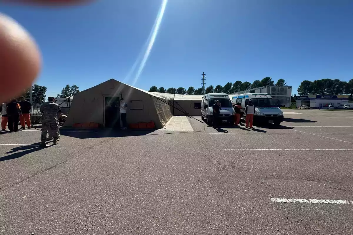 Un campamento con una carpa grande y varios vehículos estacionados en un área abierta, con personas alrededor y un cielo despejado.