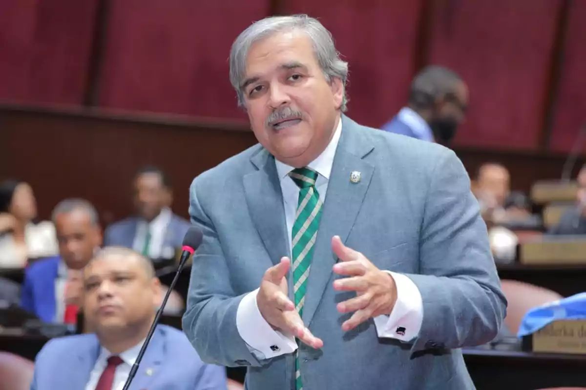 Un hombre de traje gris y corbata verde habla gesticulando en un entorno formal con personas al fondo.