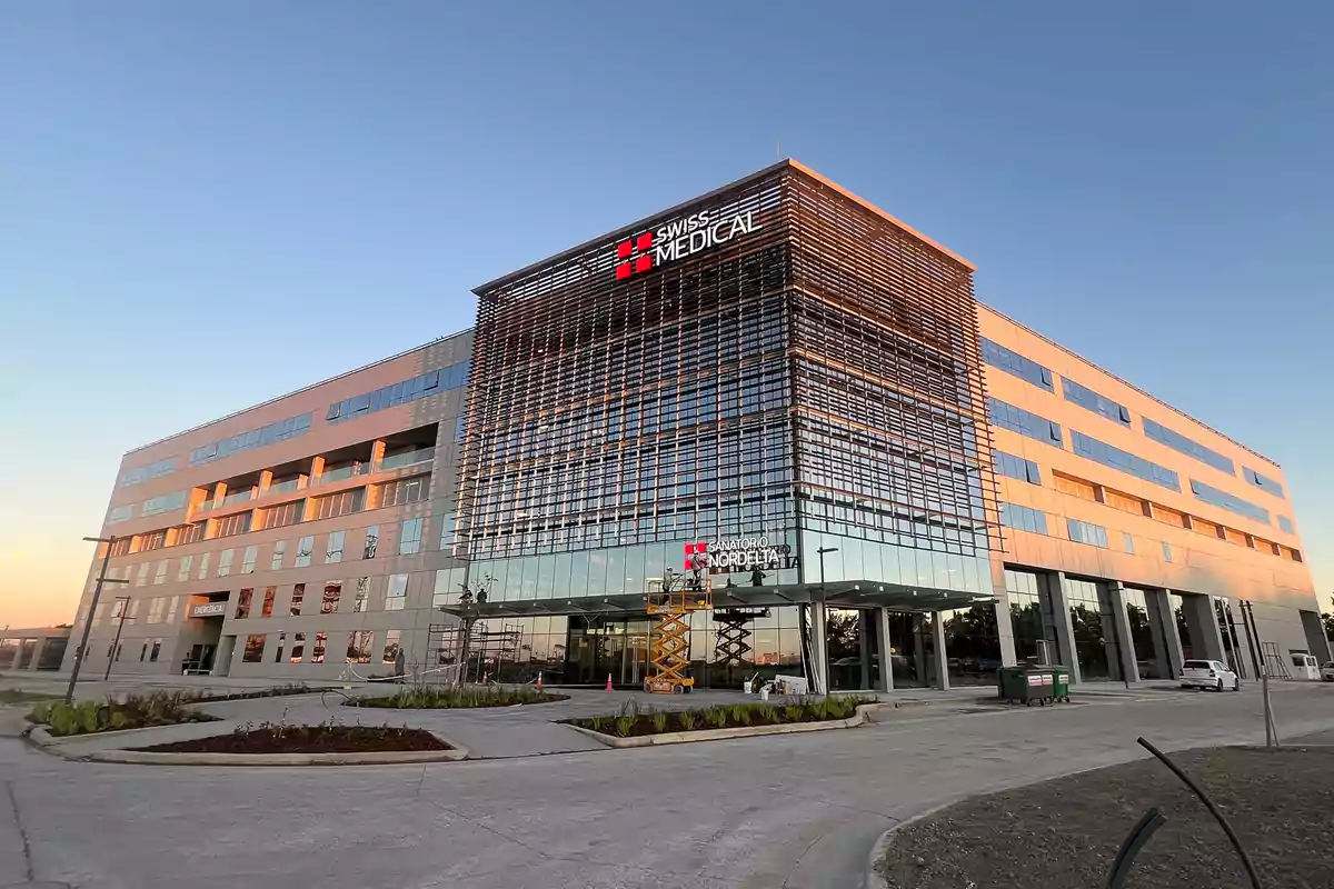 Edificio moderno de varios pisos con fachada de vidrio y el logo de Swiss Medical en la parte superior, rodeado de jardines y estacionamiento bajo un cielo despejado al atardecer