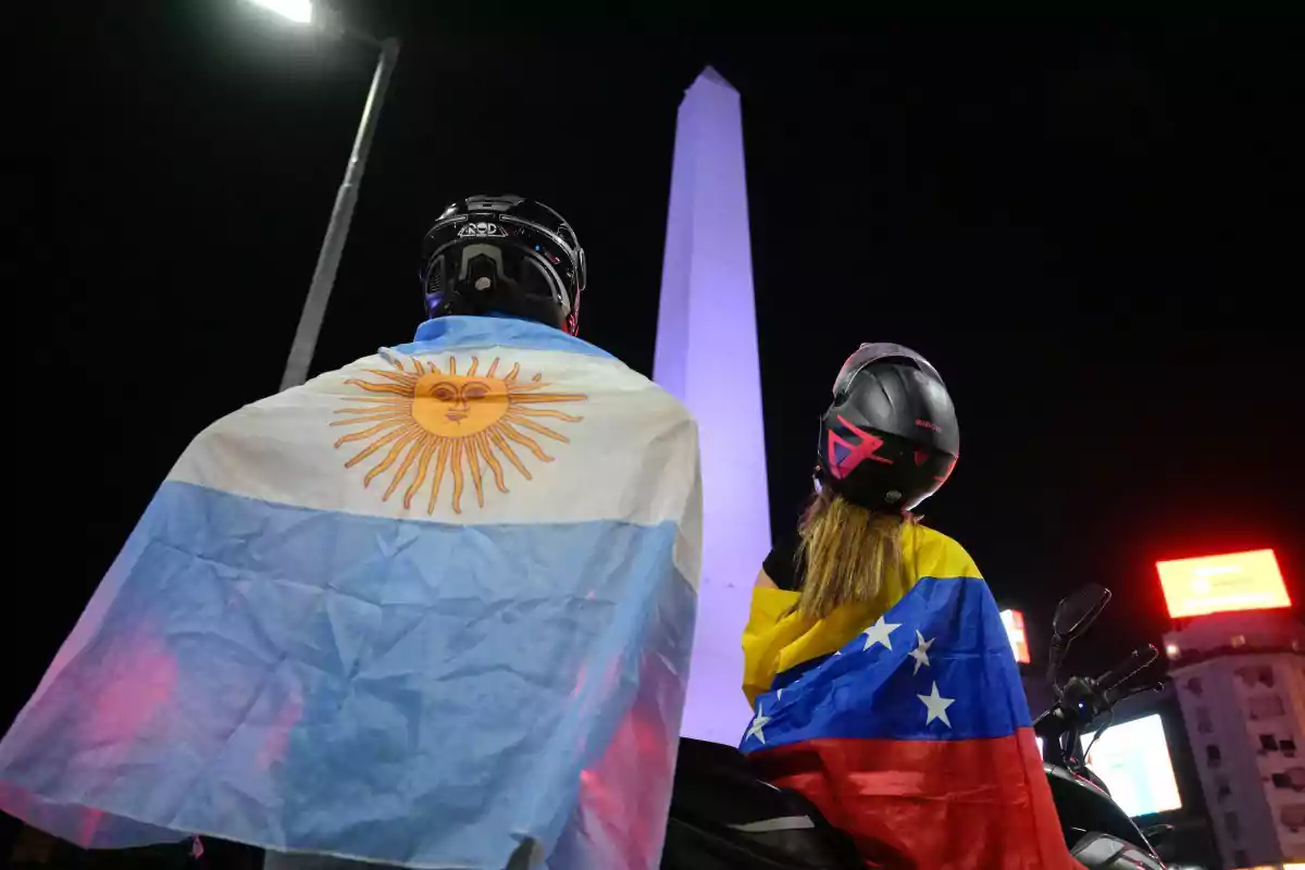 Dos personas con cascos de motocicleta llevan las banderas de Argentina y Venezuela mientras observan el Obelisco iluminado por la noche.