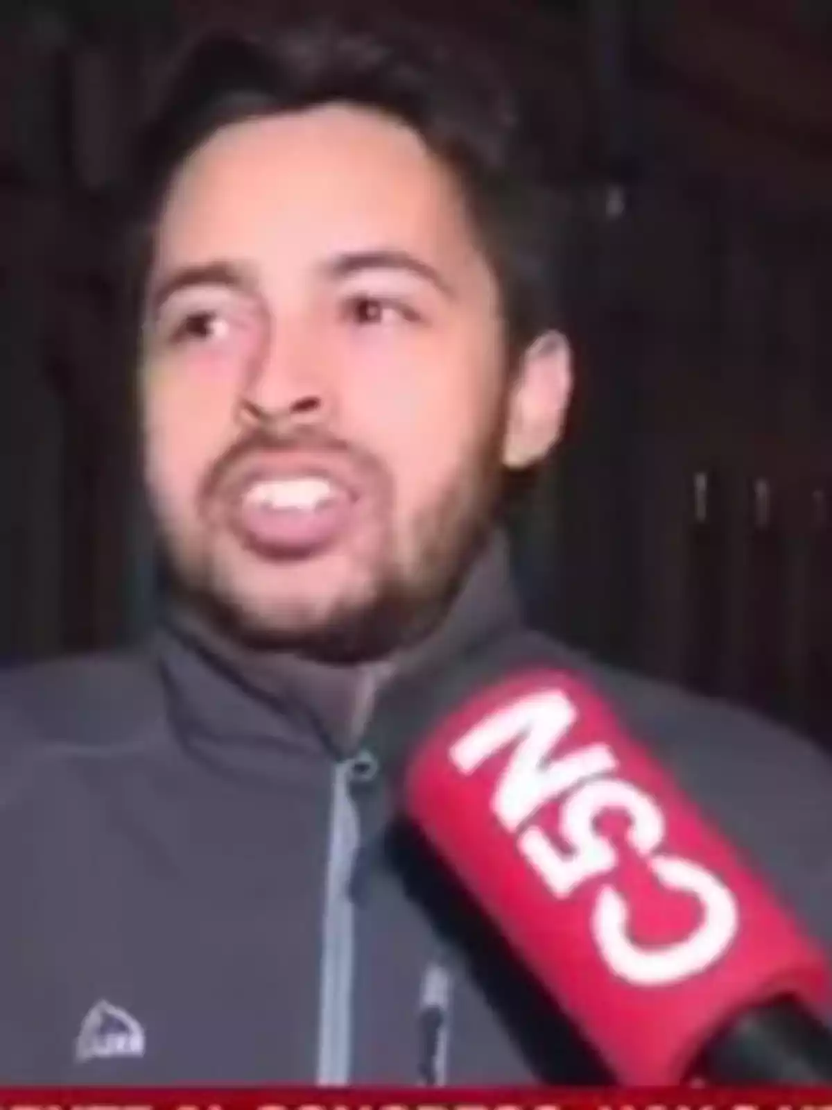 Un hombre con barba y chaqueta oscura está siendo entrevistado por un micrófono rojo de un canal de noticias.