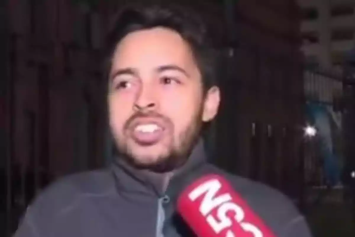 A man with a beard and a dark jacket speaks in front of a news microphone.