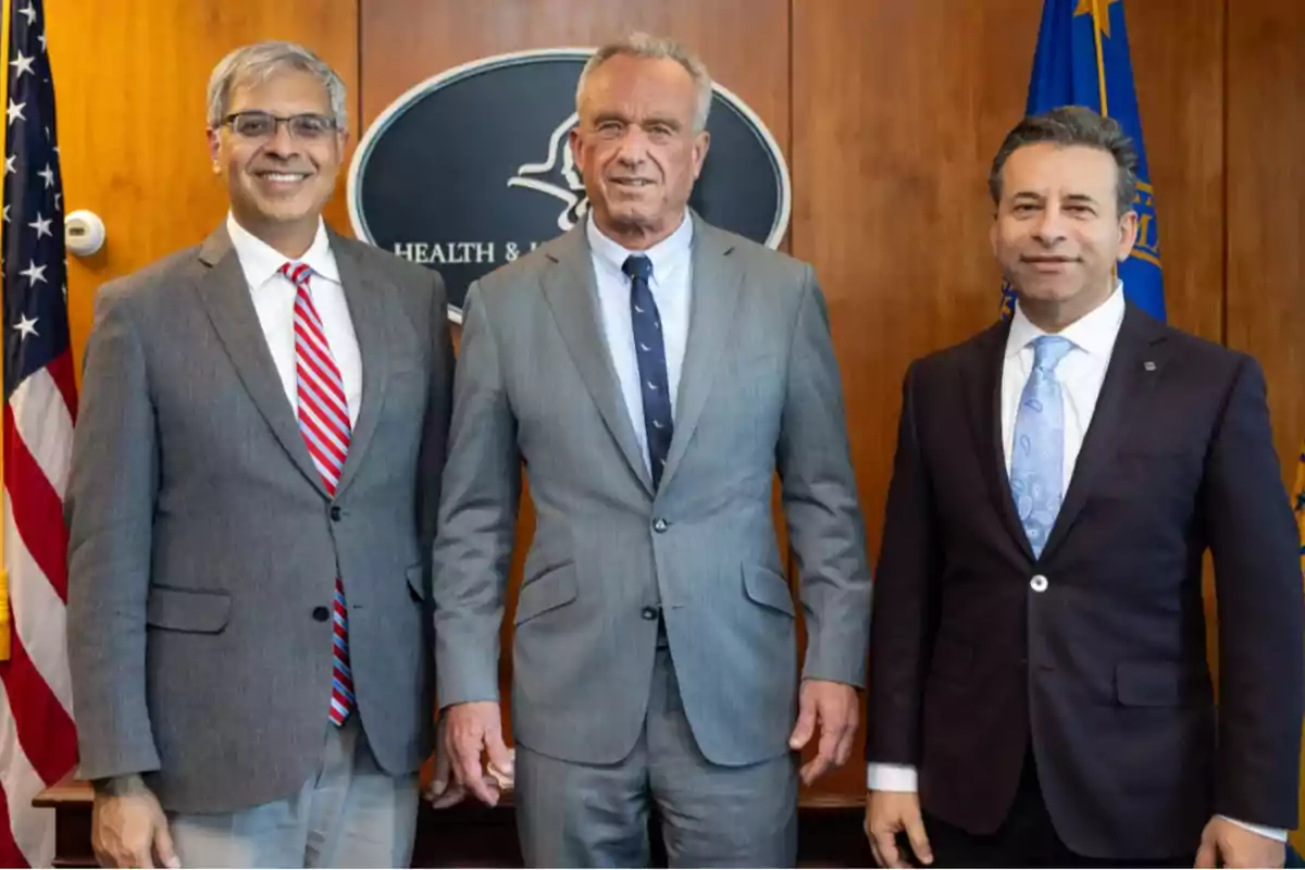 Tres hombres de traje posan juntos en una oficina con banderas y un letrero institucional al fondo Tres hombres de traje posan juntos en una oficina con banderas y un letrero institucional al fondo