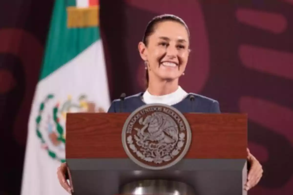 Persona sonriendo detrás de un podio con el escudo de México y una bandera mexicana al fondo.