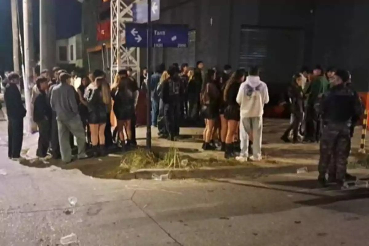 Un grupo de personas jóvenes se reúne en una esquina durante la noche cerca de un poste y un cartel de calles, algunas personas visten ropa oscura y hay un policía observando la escena.