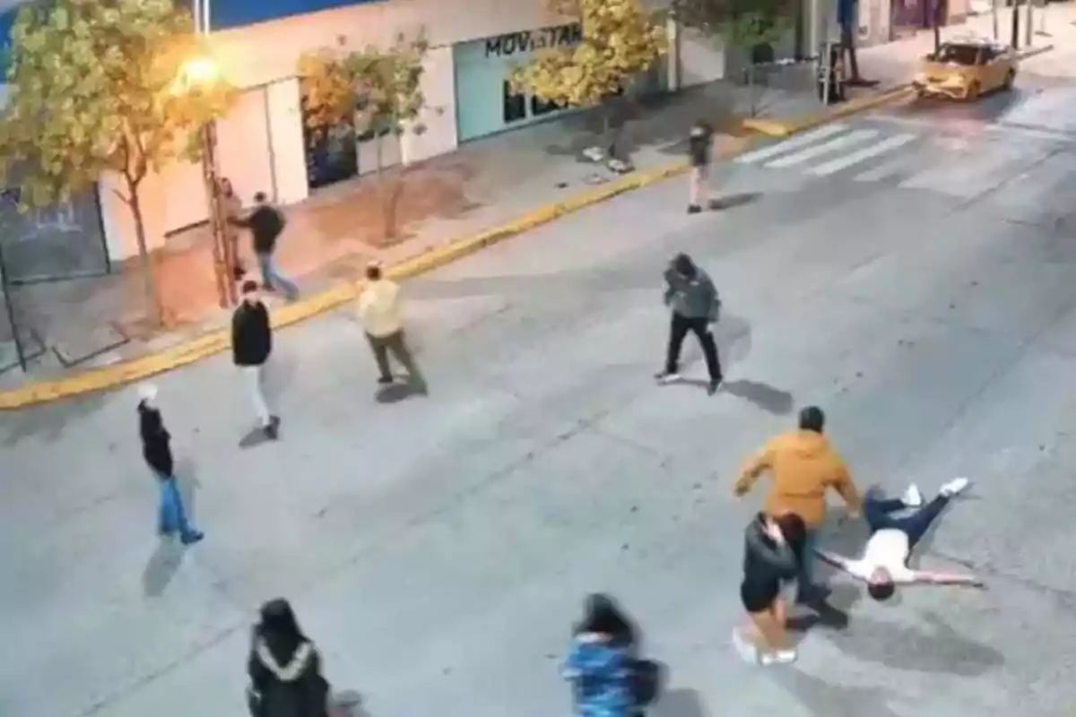 Un grupo de personas camina por una calle iluminada por farolas, mientras una persona yace en el suelo. Un grupo de personas camina por una calle iluminada por farolas, mientras una persona yace en el suelo.
