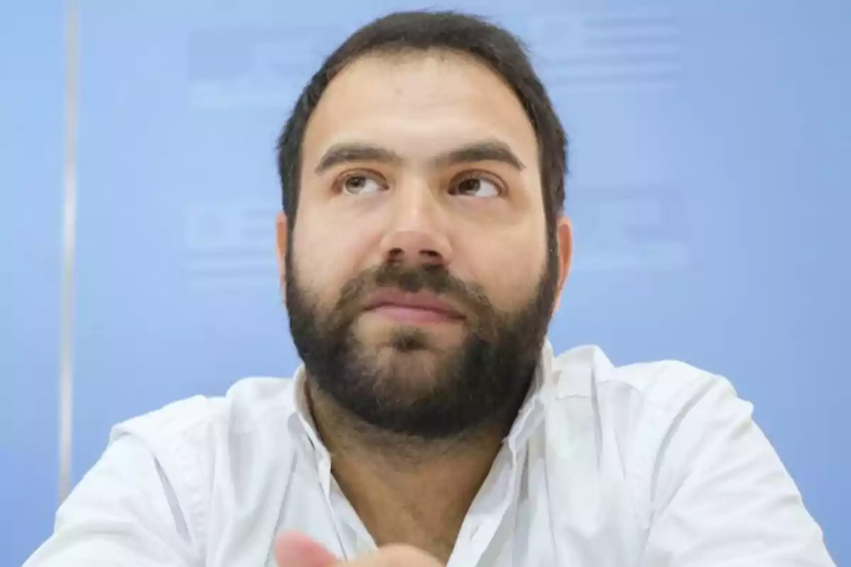 Dark-haired, bearded man in a white jersey looking up against a blue background