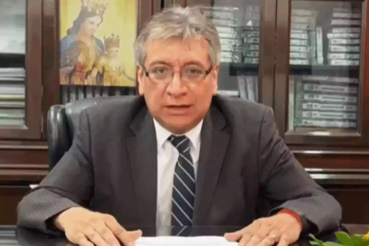 Hombre de cabello canoso y gafas con traje y corbata sentado en un escritorio con estanterías y una imagen religiosa al fondo Hombre de cabello canoso y gafas con traje y corbata sentado en un escritorio con estanterías y una imagen religiosa al fondo