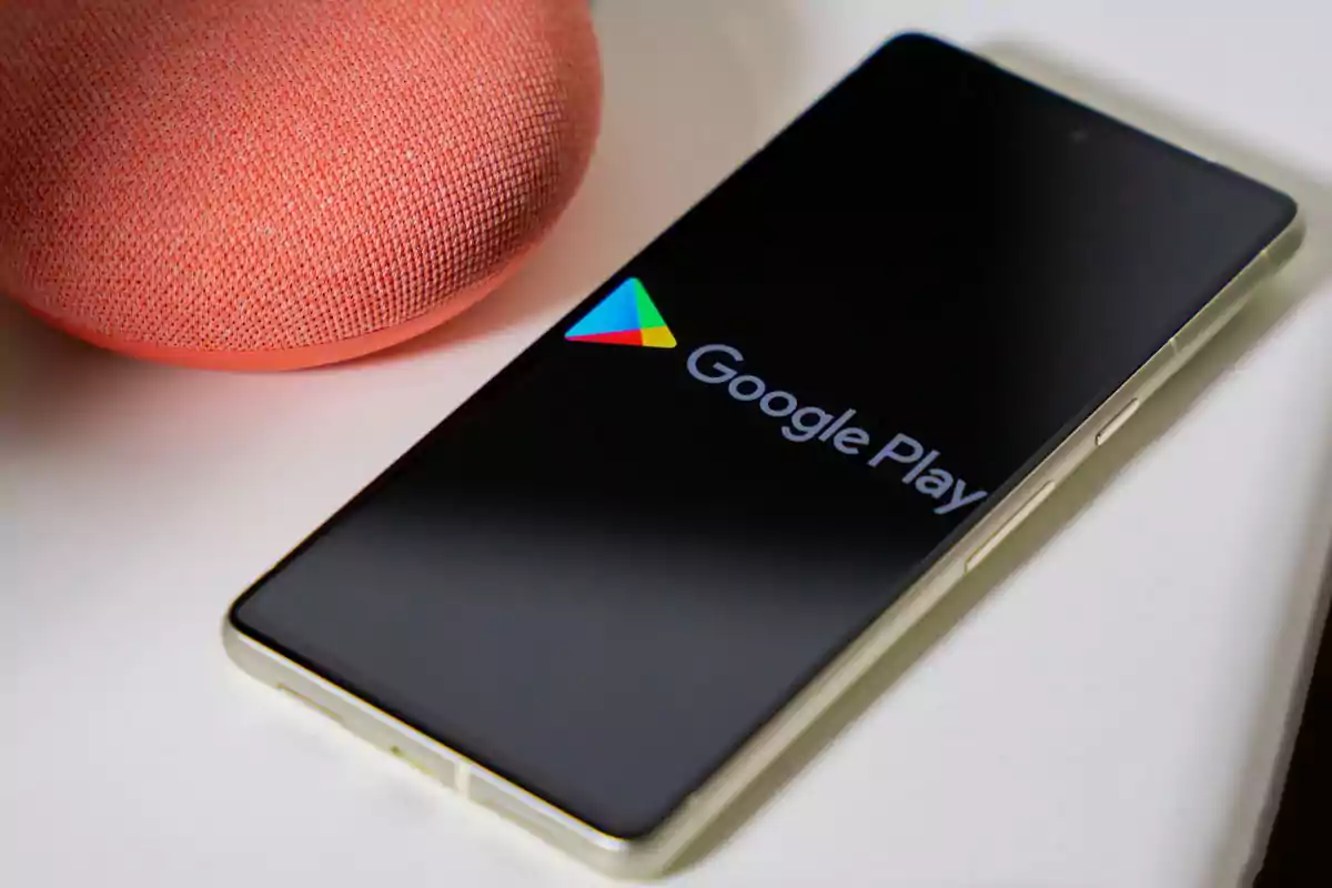 Smartphone with the screen on displaying the Google Play logo next to a coral-colored smart speaker on a white surface Smartphone with the screen on displaying the Google Play logo next to a coral-colored smart speaker on a white surface