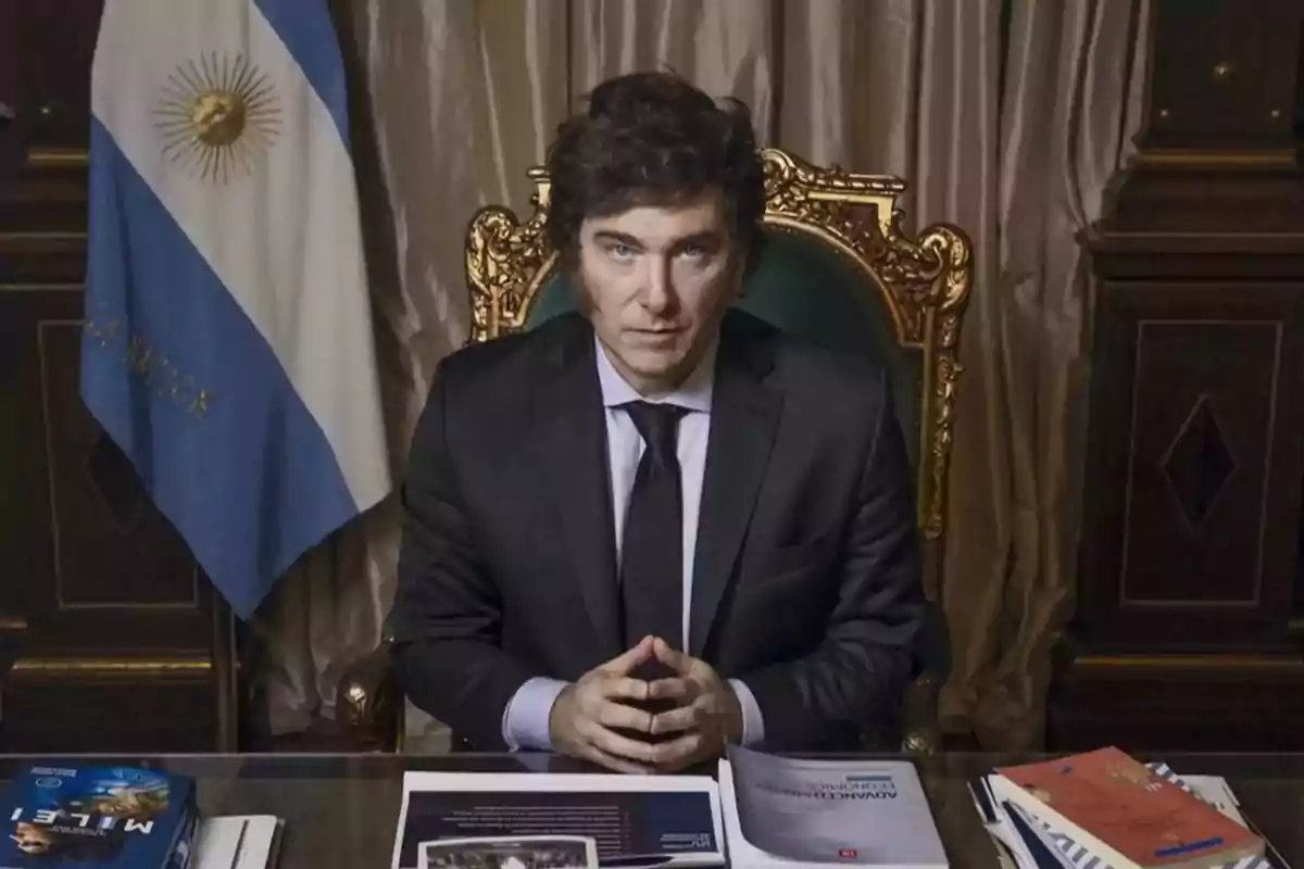 Hombre de traje sentado en un escritorio con una bandera de Argentina al fondo.