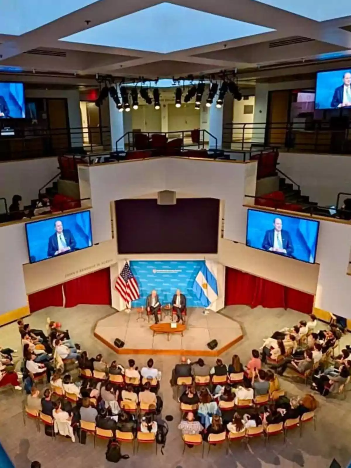 Una conferencia en un auditorio con dos personas en el escenario, rodeadas de banderas de Estados Unidos y Argentina, mientras el público está sentado observando. Una conferencia en un auditorio con dos personas en el escenario, rodeadas de banderas de Estados Unidos y Argentina, mientras el público está sentado observando.