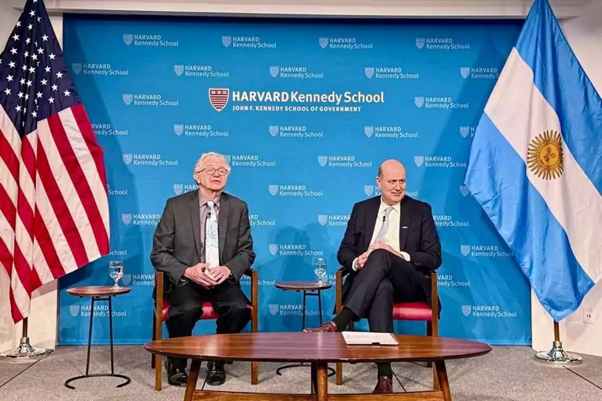 Dos hombres sentados en sillas frente a un fondo de la Harvard Kennedy School con las banderas de Estados Unidos y Argentina a los lados.