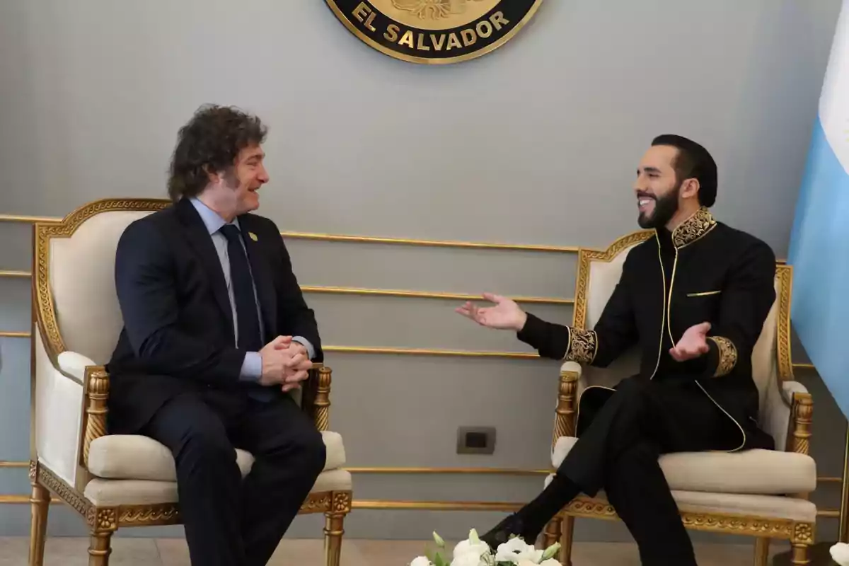 Two people sitting in golden chairs are having a lively conversation in a room with a coat of arms of El Salvador on the wall.