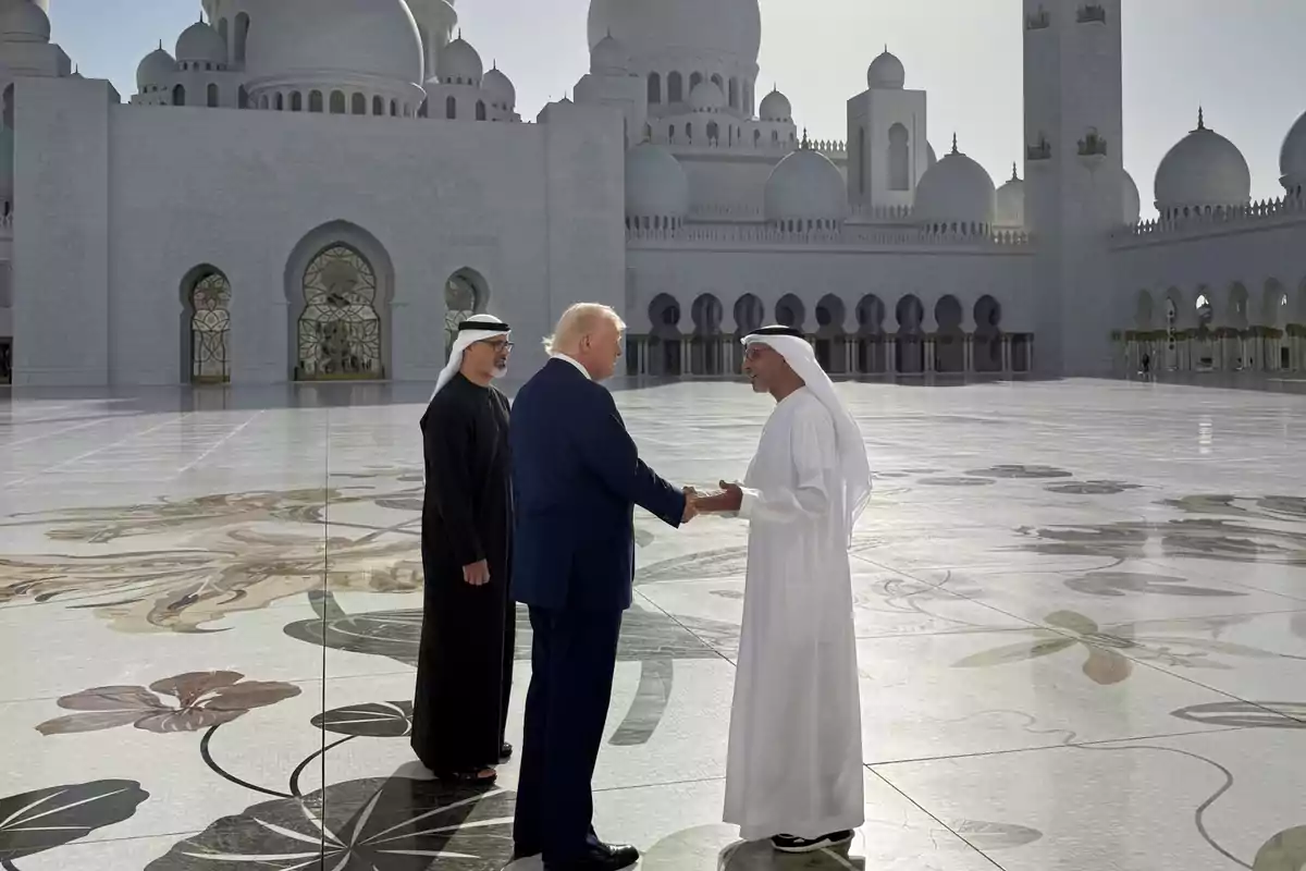 Un grupo de personas se saluda en el patio de una gran mezquita con cúpulas y arcos al fondo.