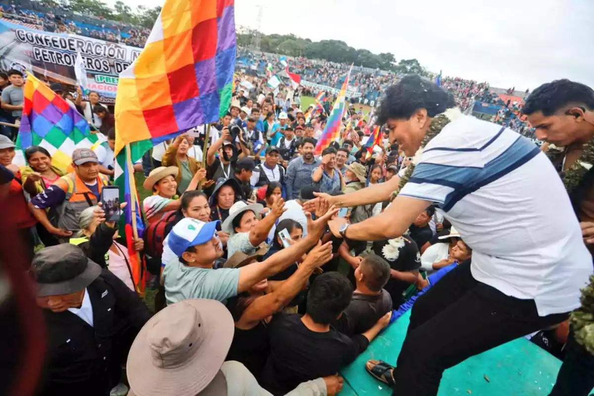 Una multitud de personas se reúne en un evento al aire libre, ondeando banderas coloridas mientras un hombre en el escenario saluda y estrecha las manos de los asistentes. Una multitud de personas se reúne en un evento al aire libre, ondeando banderas coloridas mientras un hombre en el escenario saluda y estrecha las manos de los asistentes.