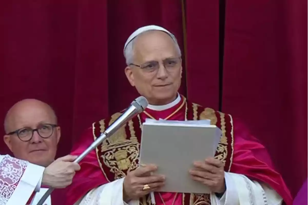 Un hombre vestido con vestimenta religiosa sostiene un micrófono y un documento frente a un fondo rojo.