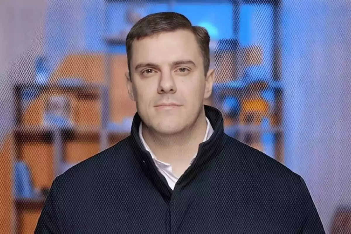 Un hombre con chaqueta oscura posando frente a un fondo desenfocado con tonos azules y naranjas.