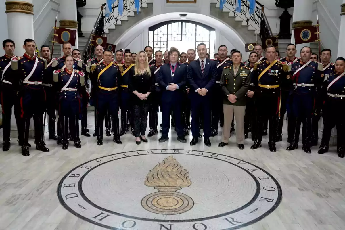 Un grupo de personas, algunas en uniformes militares, posan en un edificio con escaleras y banderas en el fondo, sobre un piso con un emblema que dice "Deber Libertad Valor Honor".