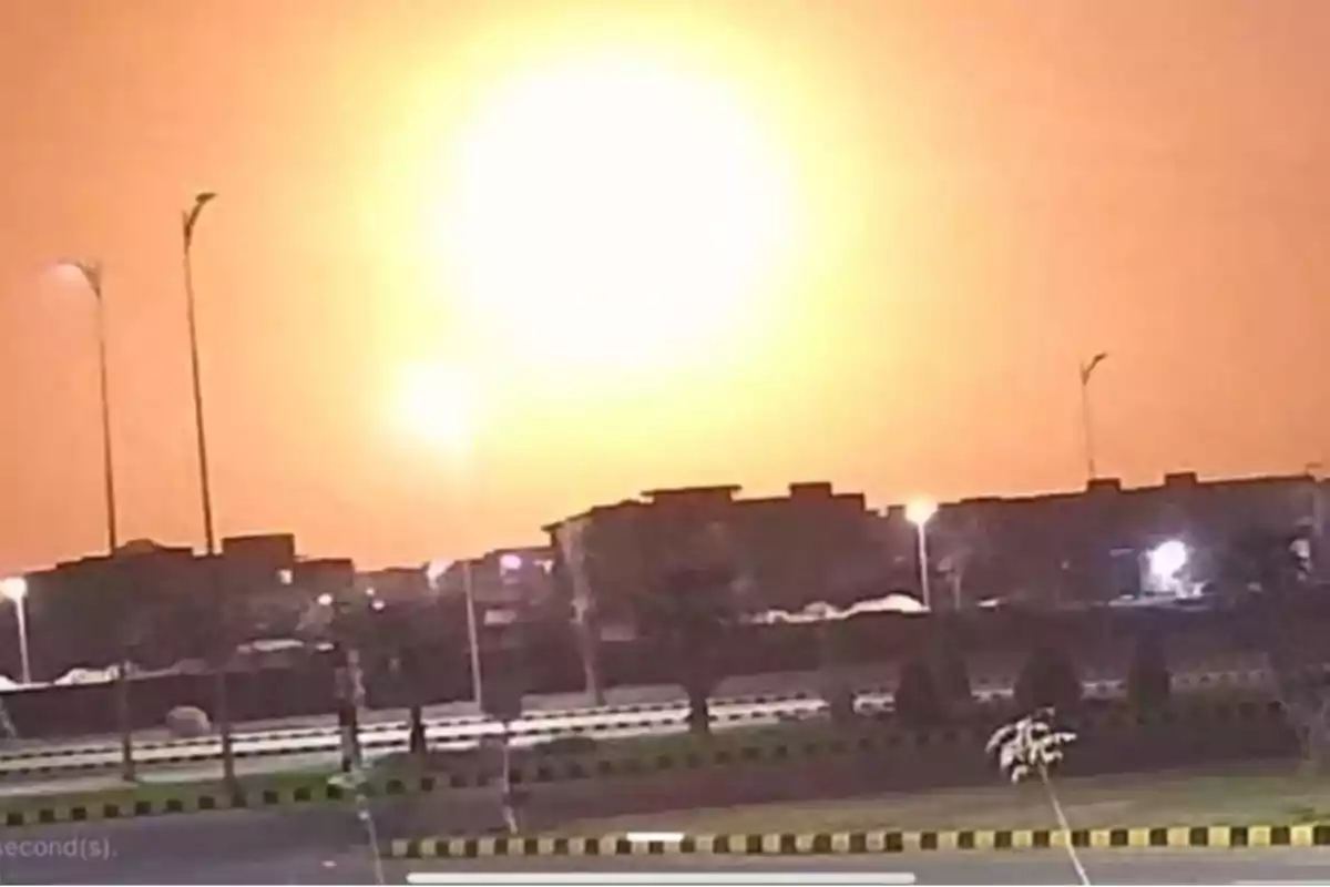 Una explosión brillante ilumina el cielo nocturno sobre una ciudad con edificios y farolas en primer plano.