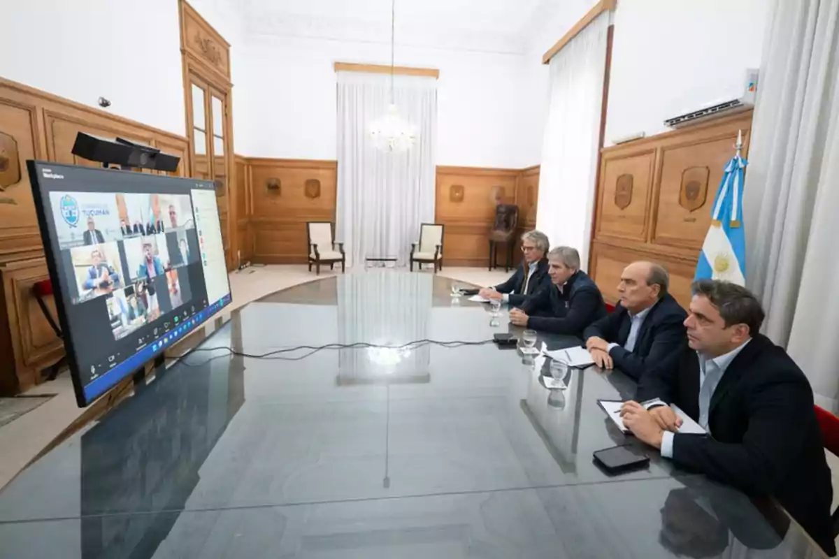 Four men sitting at a glass table participate in a video call in an elegant room with wooden walls and an Argentine flag.