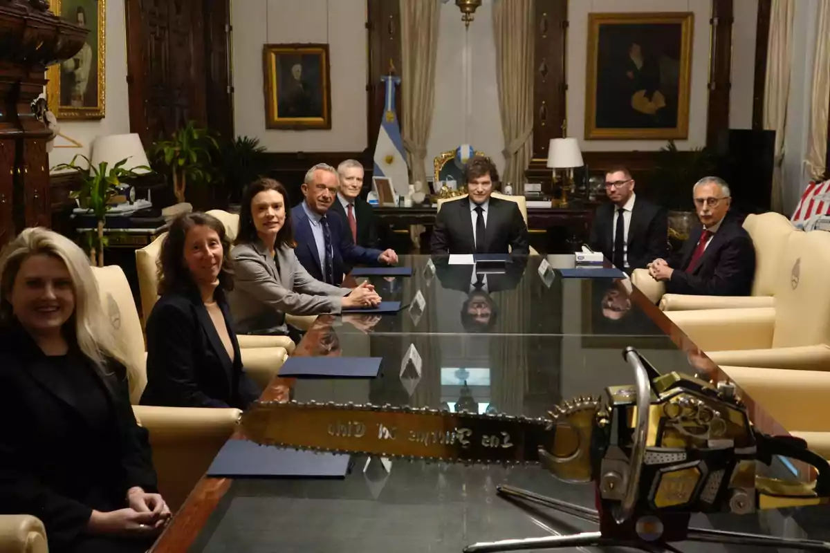 Ocho personas sentadas alrededor de una mesa de reuniones en una oficina elegante con una motosierra dorada sobre la mesa y la bandera de Argentina al fondo