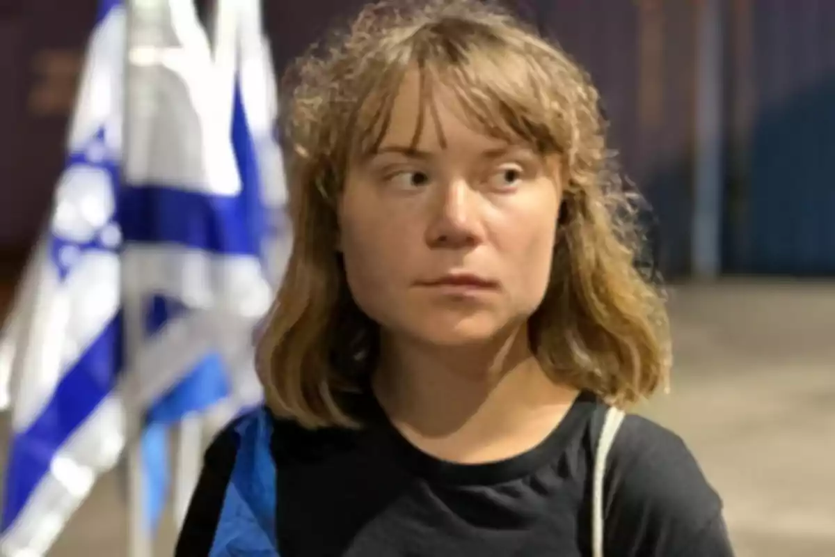 Joven de cabello rubio y corto con expresión seria, viste camiseta negra y está de pie frente a una bandera azul y blanca desenfocada en el fondo Joven de cabello rubio y corto con expresión seria, viste camiseta negra y está de pie frente a una bandera azul y blanca desenfocada en el fondo