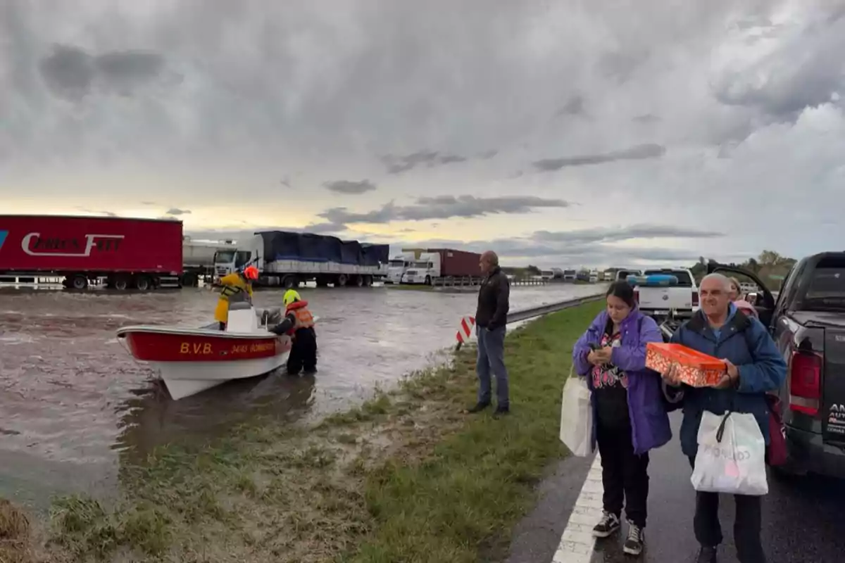 People watch as rescuers in a boat navigate a flooded road, with stalled trucks in the background and a cloudy sky.