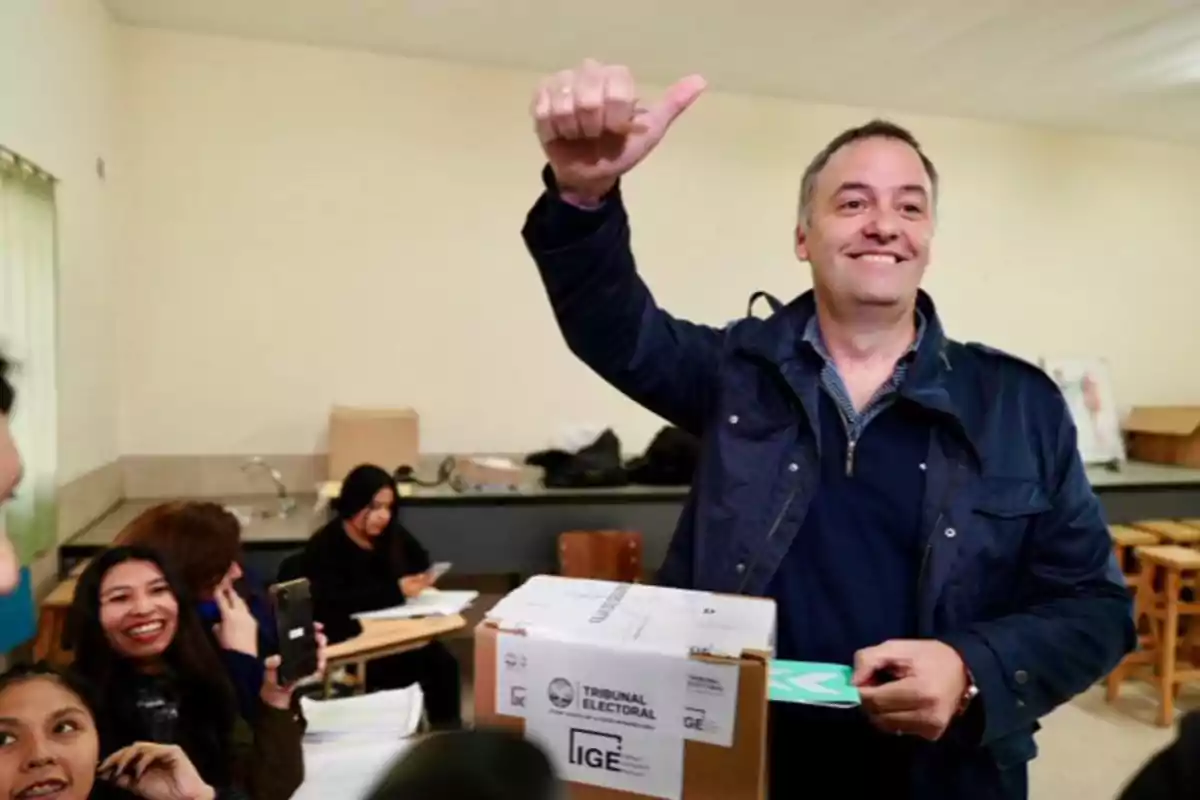 Un hombre sonriente levanta el pulgar mientras vota en una urna electoral, rodeado de personas que observan y sonríen.