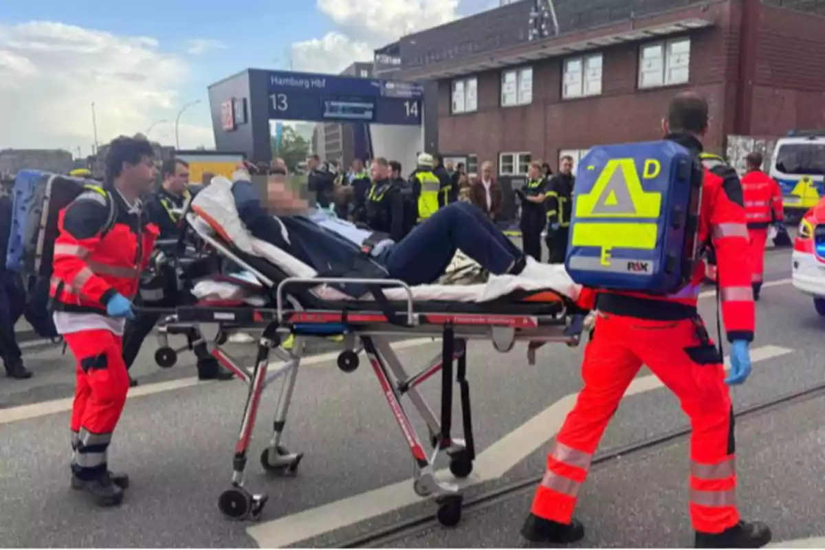 A group of paramedics is transporting a person on a stretcher near a train station with several emergency vehicles and security personnel present at the scene. A group of paramedics is transporting a person on a stretcher near a train station with several emergency vehicles and security personnel present at the scene.