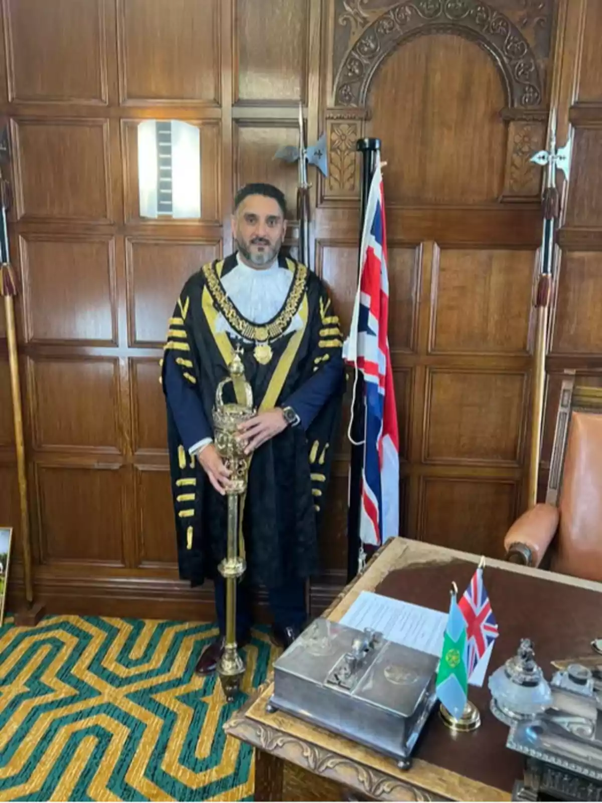 A man dressed in a ceremonial robe and a golden chain stands next to a United Kingdom flag in a wood-paneled room, holding a golden scepter next to a desk with documents and small flags. A man dressed in a ceremonial robe and a golden chain stands next to a United Kingdom flag in a wood-paneled room, holding a golden scepter next to a desk with documents and small flags.