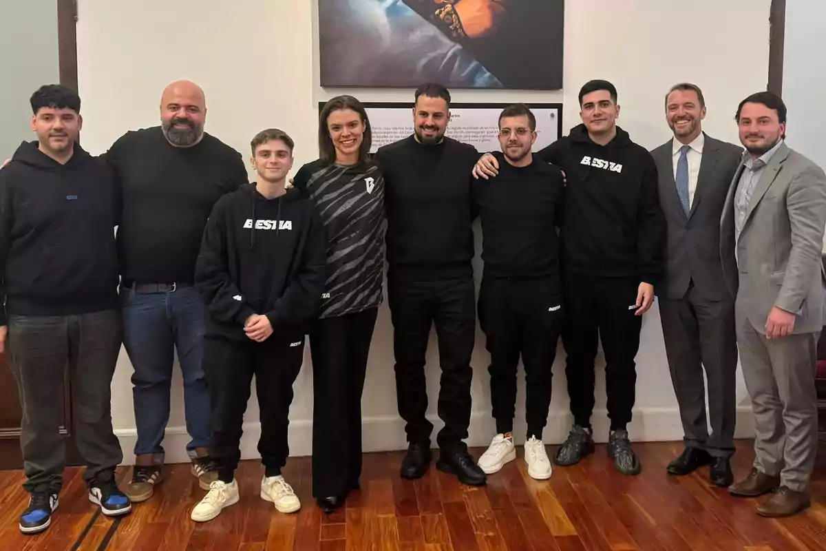 Un grupo de personas posando juntas en una habitación con suelo de madera, algunas visten ropa casual y otras trajes formales, y hay un cuadro colgado en la pared detrás de ellos.
