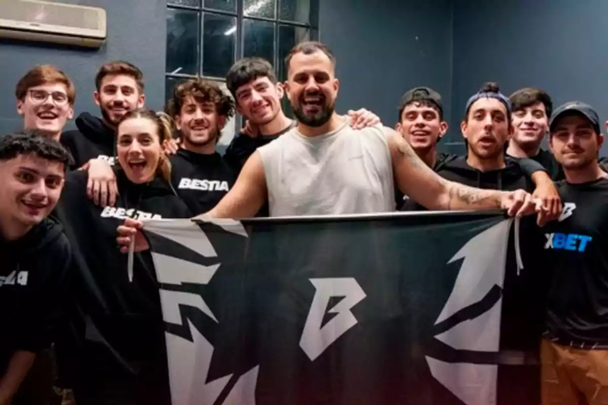Un grupo de personas sonrientes posando juntas, algunas de ellas llevan sudaderas con la palabra "BESTIA" y sostienen una bandera con un diseño gráfico.