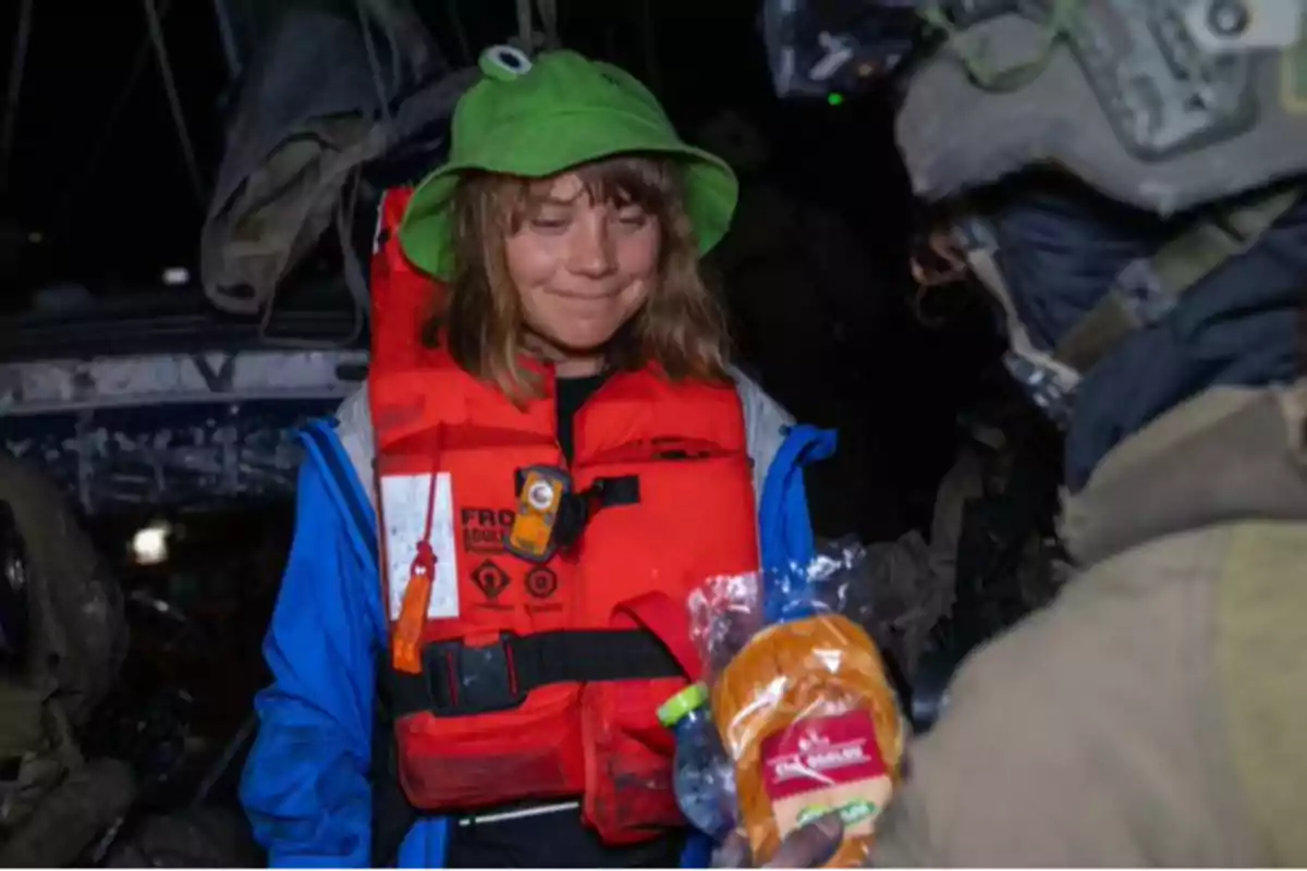 Woman wearing a red life jacket and a green hat smiles as she receives a loaf of bread from a person dressed in a military uniform.