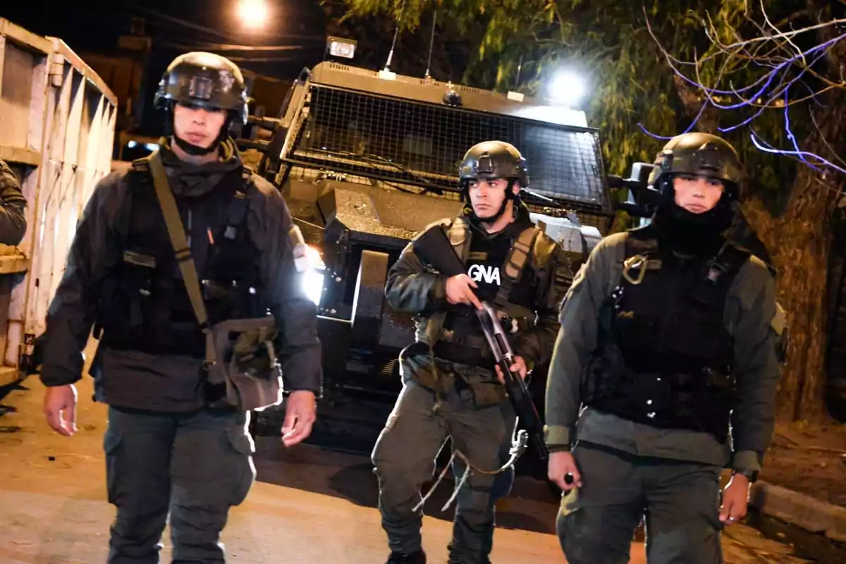 Tres agentes de seguridad armados y equipados con chalecos antibalas y cascos, caminando frente a un vehículo blindado en una operación nocturna.