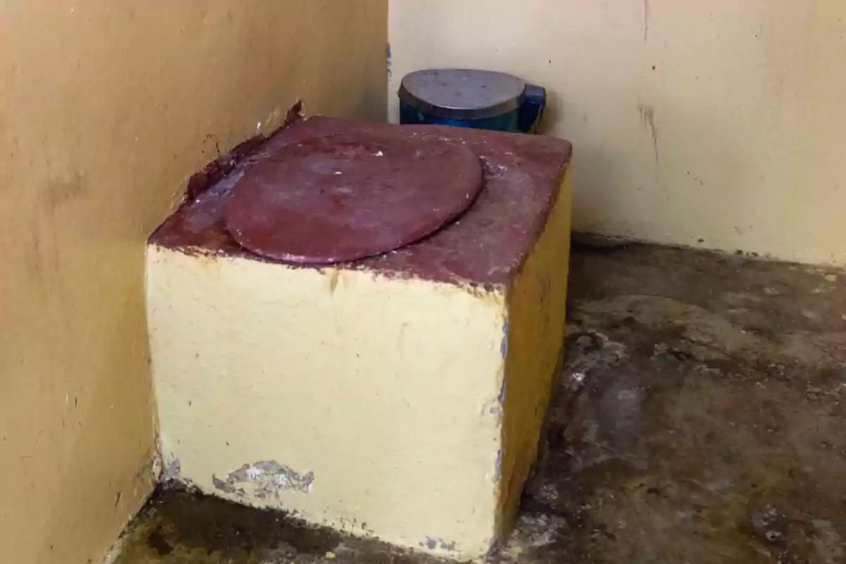 Baño rústico de letrina de cemento con tapa circular en una esquina de paredes amarillas y piso de concreto