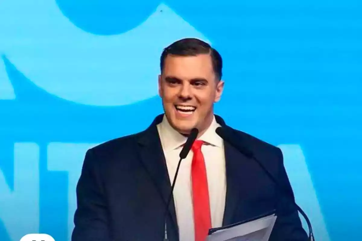 Short dark-haired man wearing a suit and red tie, smiling as he speaks in front of two microphones with a blue background